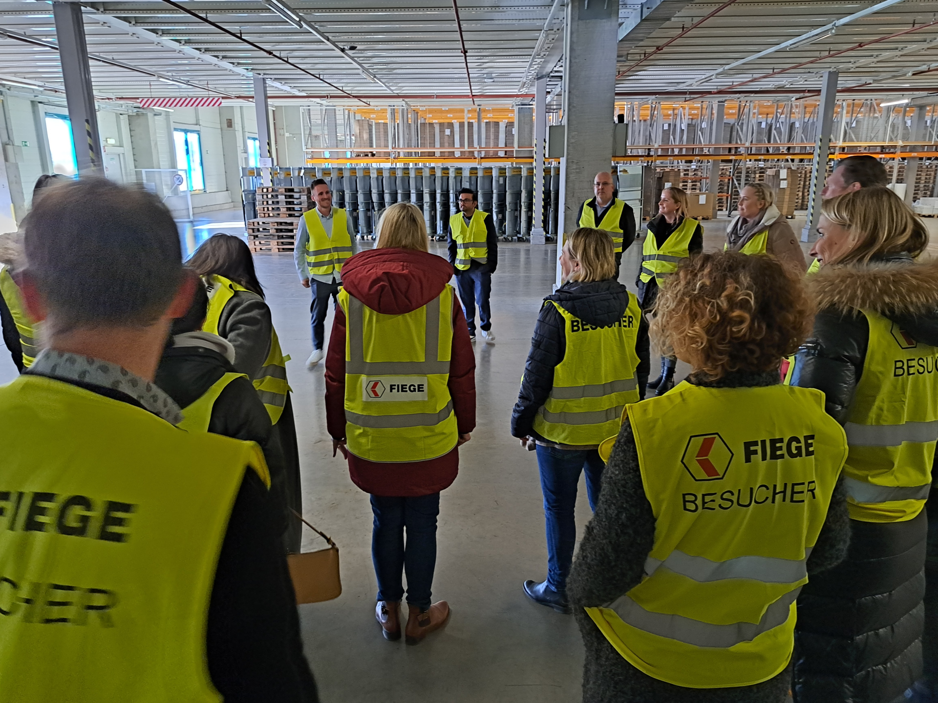 Eine Gruppe von Menschen in gelben Warnwesten steht in einer Lagehalle in einem Kreis.